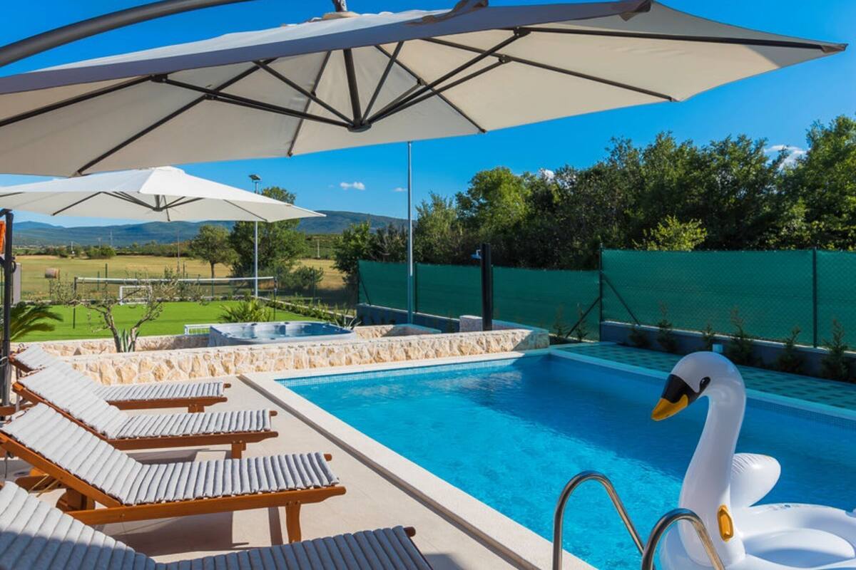 A swimming pool with lounge chairs and umbrellas, surrounded by a green fence and trees.