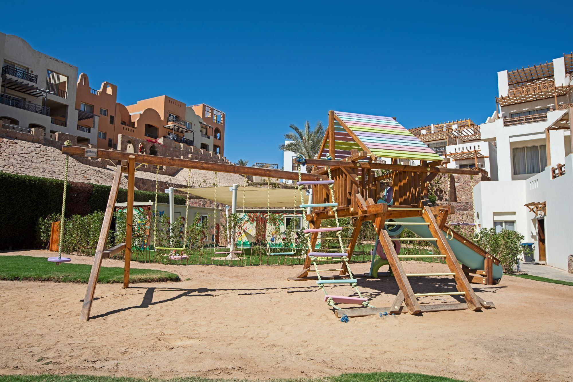 Un terrain de jeux avec une structure d’escalade en bois et des balançoires, situé dans un espace sablonneux, entouré d’immeubles dotés de balcons, le tout sous un ciel bleu dégagé.