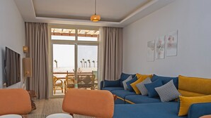 A dining area with a table set for a meal, a blue sofa with yellow cushions, and a view of the beach through the window. - Spacious Two bedroom in Azzurra with Terrace Sahl Hasheesh (Sahl)