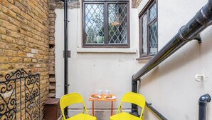 A narrow outdoor space with a brick wall, a window with a diamond pattern, and a small table with chairs. - Evershot Rd N4, Crouch Hill, Finsbury Park (London)