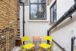 A narrow outdoor space with a brick wall, a window with a diamond pattern, and a small table with chairs.