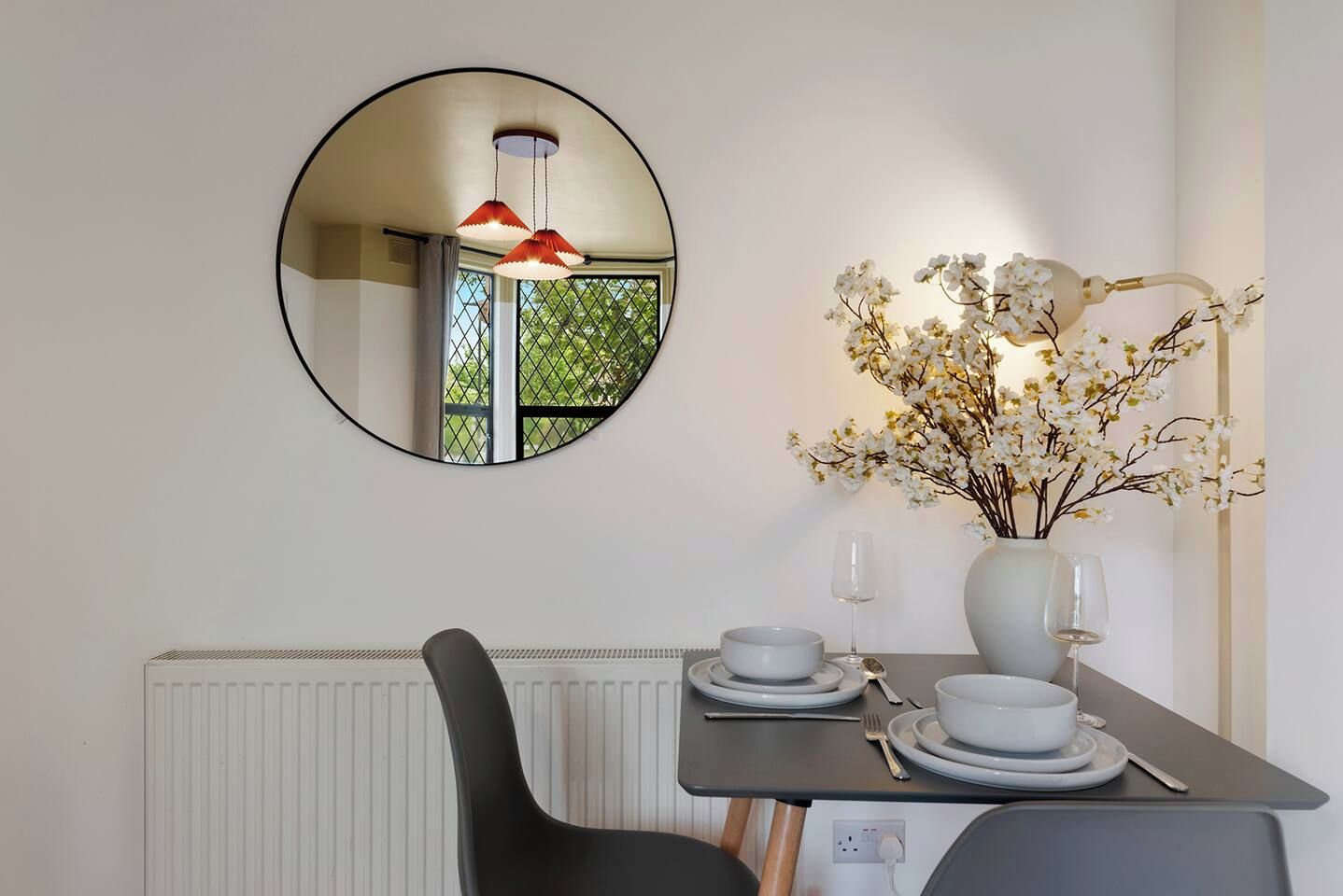 A dining area with a round mirror, a vase with flowers, and a table set for two.