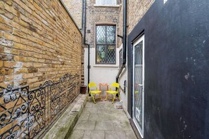 A narrow alleyway with a brick wall on one side and a black wall on the other. There are two yellow chairs and a small table in the center.