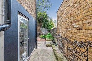 A narrow alleyway with a black metal fence, a brick wall, and a small garden area with plants and a chair.