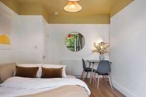 A bedroom with a bed, a desk with a chair, a round mirror, and a vase with flowers.