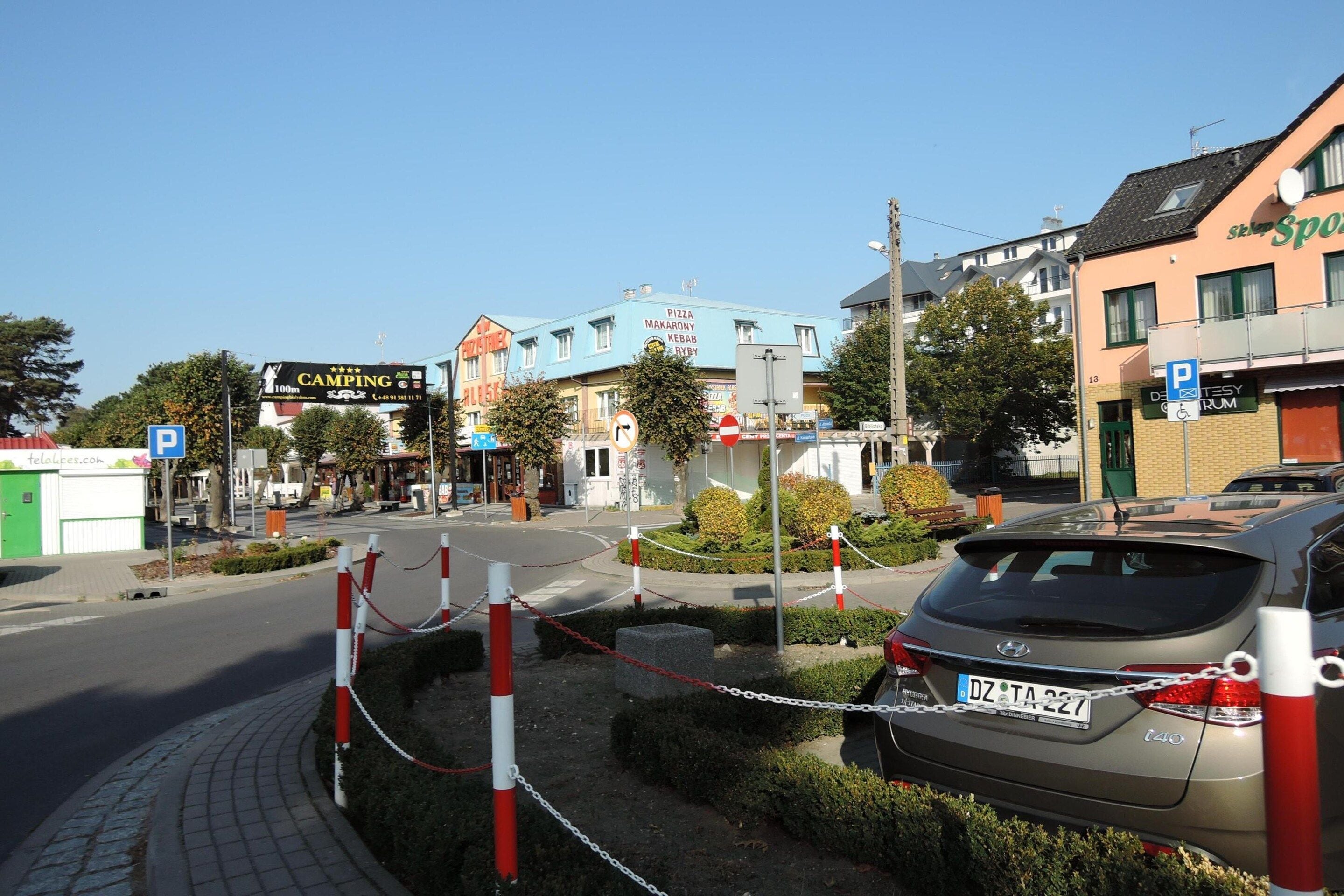 A street view with a car parked on the side, a blue sign with a white 'P' indicating parking, and a building with a sign that reads 'CAMPING'.
