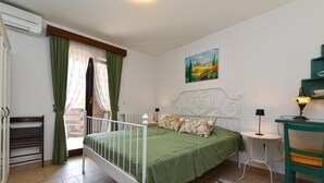 A bedroom with a bed, a small table, a chair, a radiator, a painting on the wall, and a window with curtains.