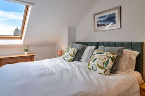 A neatly made bed with white bedding and patterned pillows, a bedside table with a lamp, a framed picture on the wall, and a window with a view of the sky and a building.