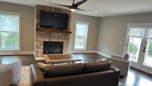 A living room with a stone fireplace, a flat-screen TV, a ceiling fan, and large windows.