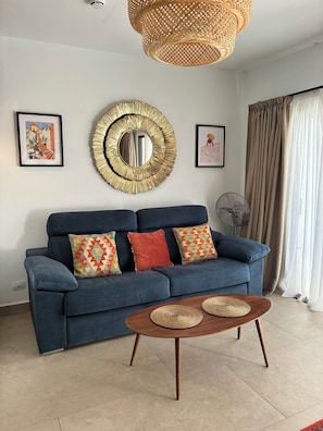 A living room with a blue sofa, two framed pictures, a round mirror, a woven light fixture, and a fan.