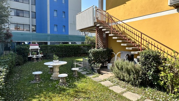 A courtyard with a small garden, a staircase, and a building with a blue and yellow facade. - Appartamento Casa Delle Rose (San Michele al Tagliamento)