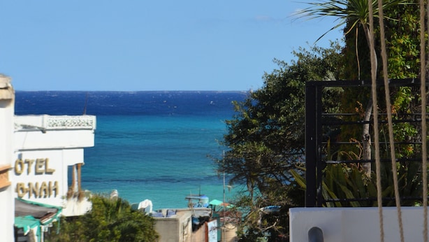 A coastal street with a view of the sea, a hotel named "HOTEL SHAH" on the left, and a narrow alley with buildings and trees. - Calma Chicha Rooms (Playa del Carmen)
