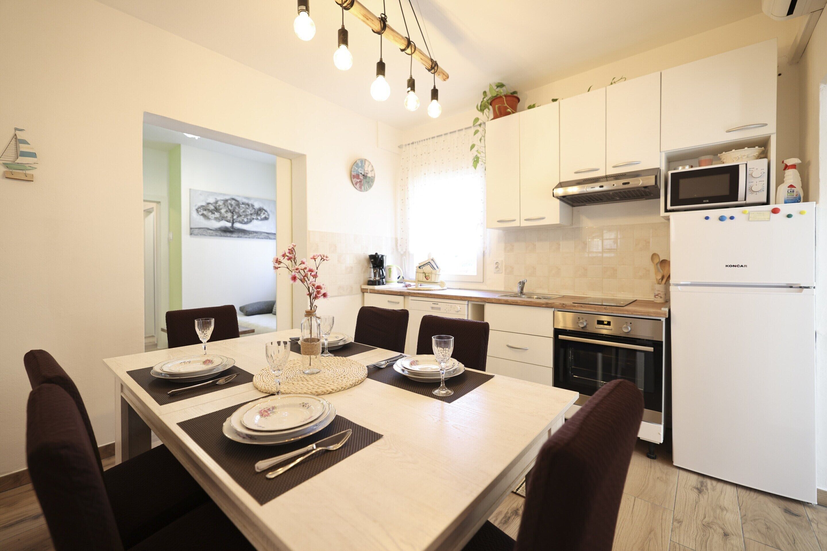 Una cocina moderna con armarios blancos, microondas, horno y refrigerador. Hay una mesa de comedor dispuesta con platos, vasos y cubiertos.