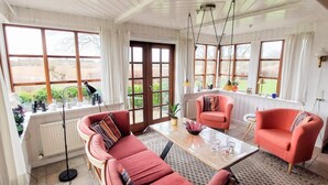 Una sala de estar acogedora con un sofá rojo, un sillón a juego y una mesita de centro. Cuenta con ventanas grandes con cortinas, un radiador y una lámpara.