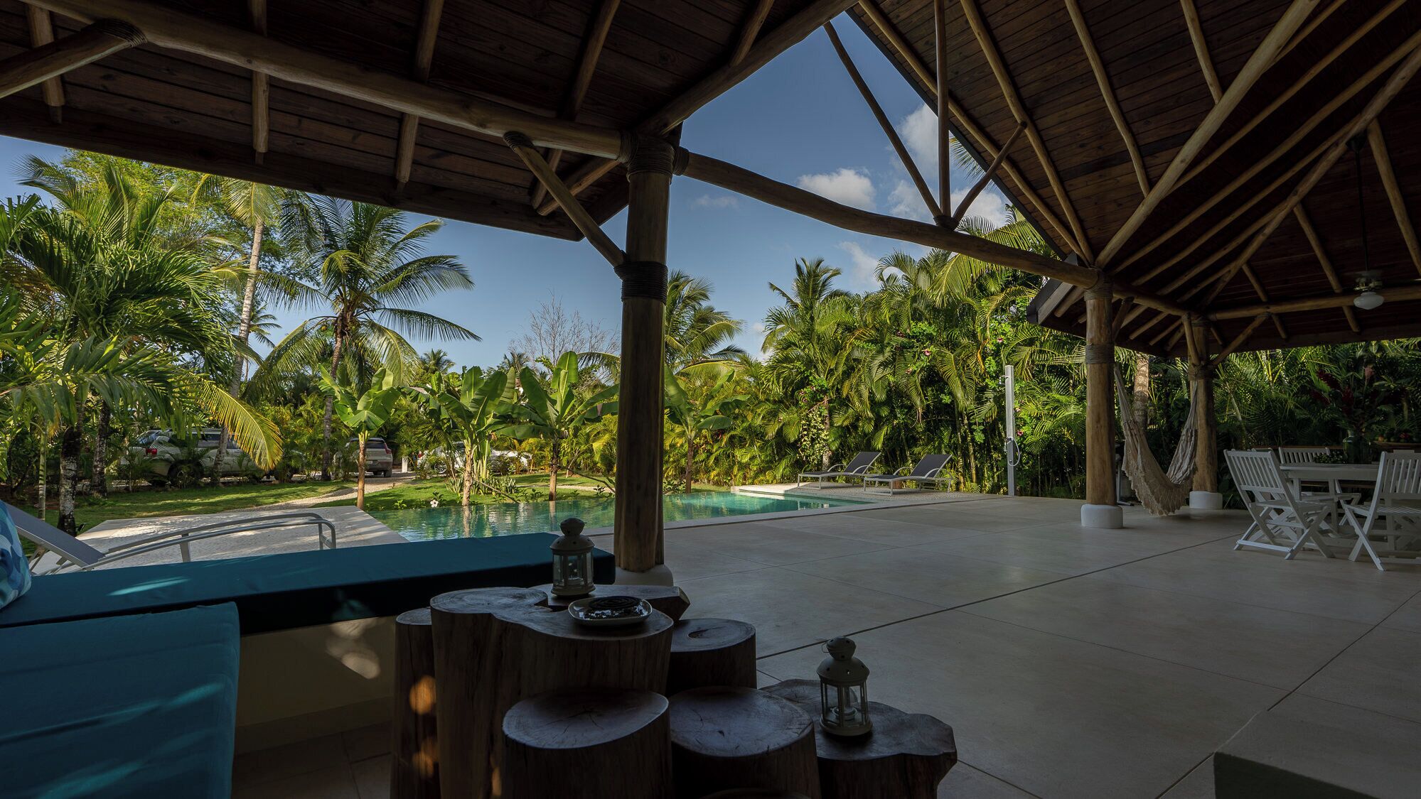 A covered outdoor area with a pool, wooden furniture, and tropical vegetation.