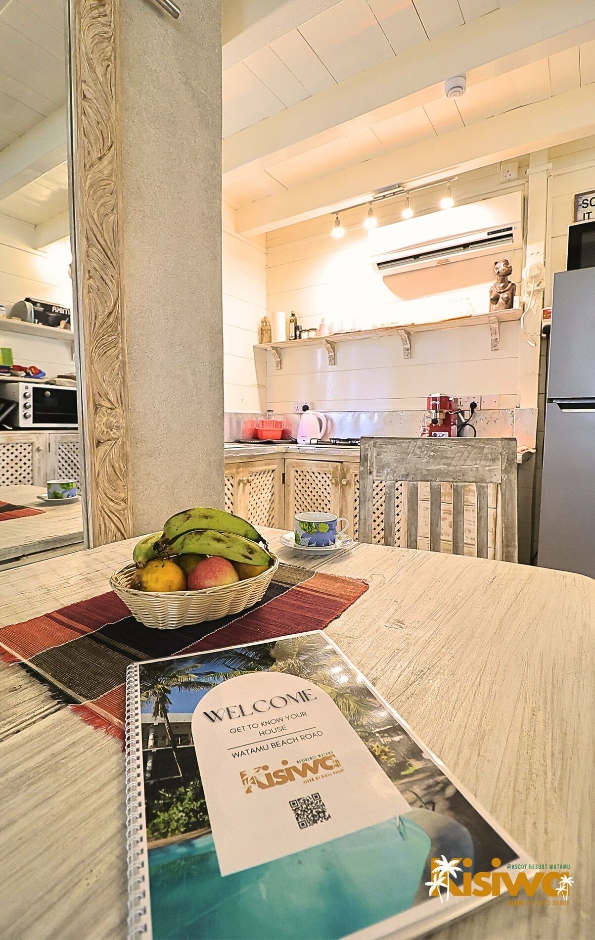 A kitchen with a fruit basket on the table, a welcome sign, and a view of the beach.