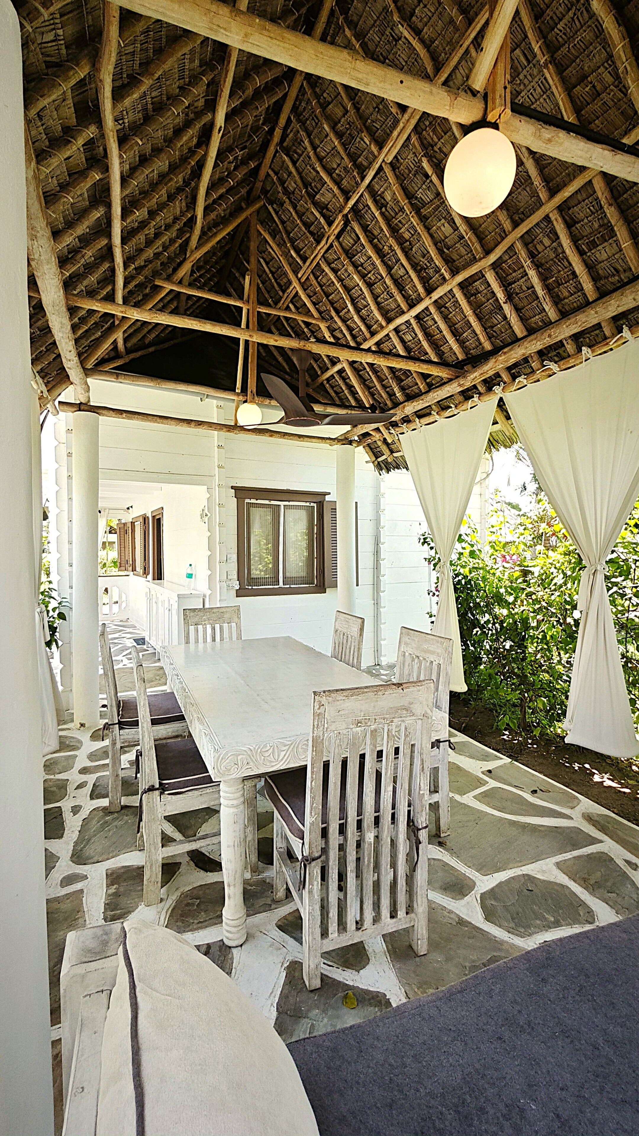 An outdoor dining area with a thatched roof, stone flooring, and white furniture.