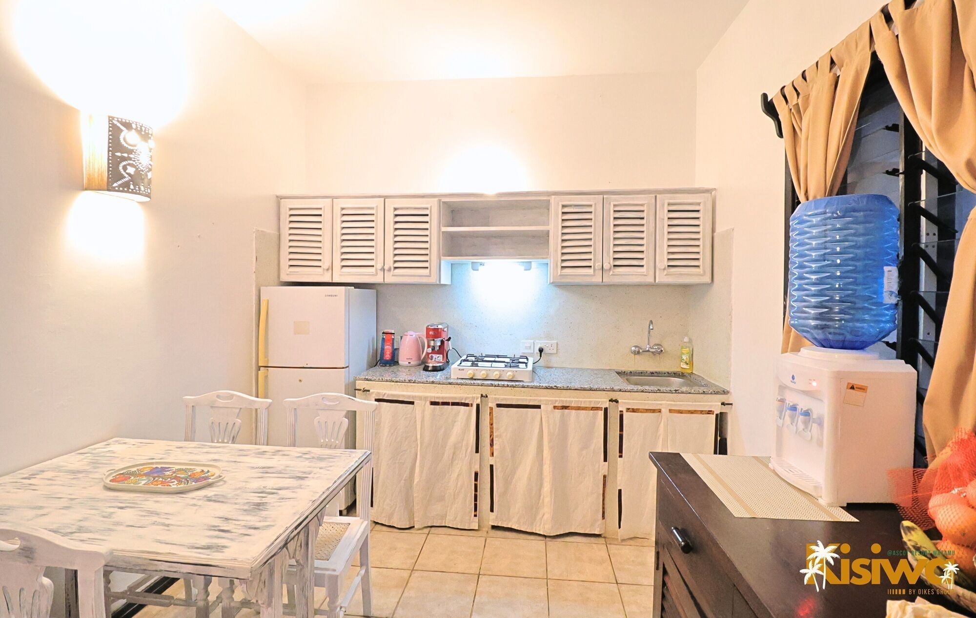 A compact kitchen with white cabinets, a refrigerator, a gas stove, and a sink. There is a dining table with chairs and a water dispenser.