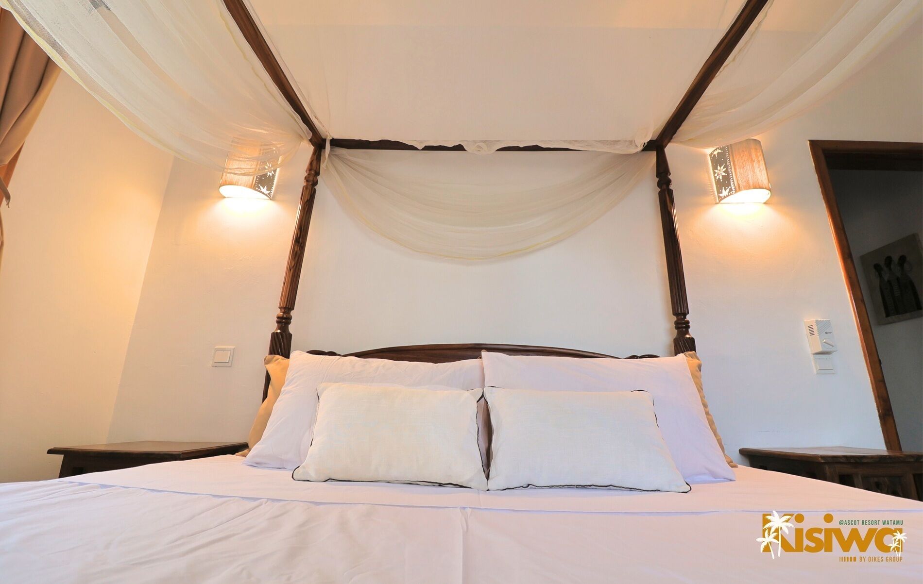 Una cama con dosel, ropa de cama y almohadas blancas, y dos luces fijadas a la pared.