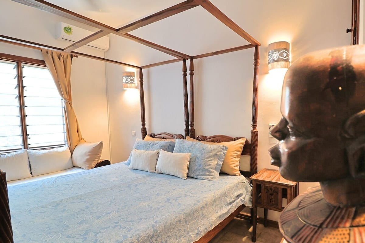 Un lit à baldaquin avec une couvre-lit à motifs, une table de chevet avec une lampe et une sculpture décorative en forme de tête.