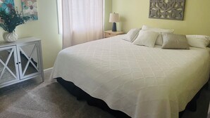 A neatly arranged bedroom with a large bed, a bedside table with a lamp, a decorative wall piece, and a window with curtains. - Charming 2-bedroom cottage with WiFi, AC in fabulous Elk Rapids (Elk Rapids)