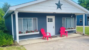 A small, single-story building with a white exterior and a dark blue roof. It features a white door, two windows with white curtains, and a large star decoration above the door. There are two red plastic chairs placed outside on the porch. - Charming 2-bedroom cottage with WiFi, AC in fabulous Elk Rapids (Elk Rapids)