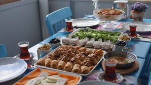 A table set with various dishes, including a tray of pastries, a bowl of olives, and a plate of sliced vegetables. - ADA LUNA BOZCAADA (Bozcaada)