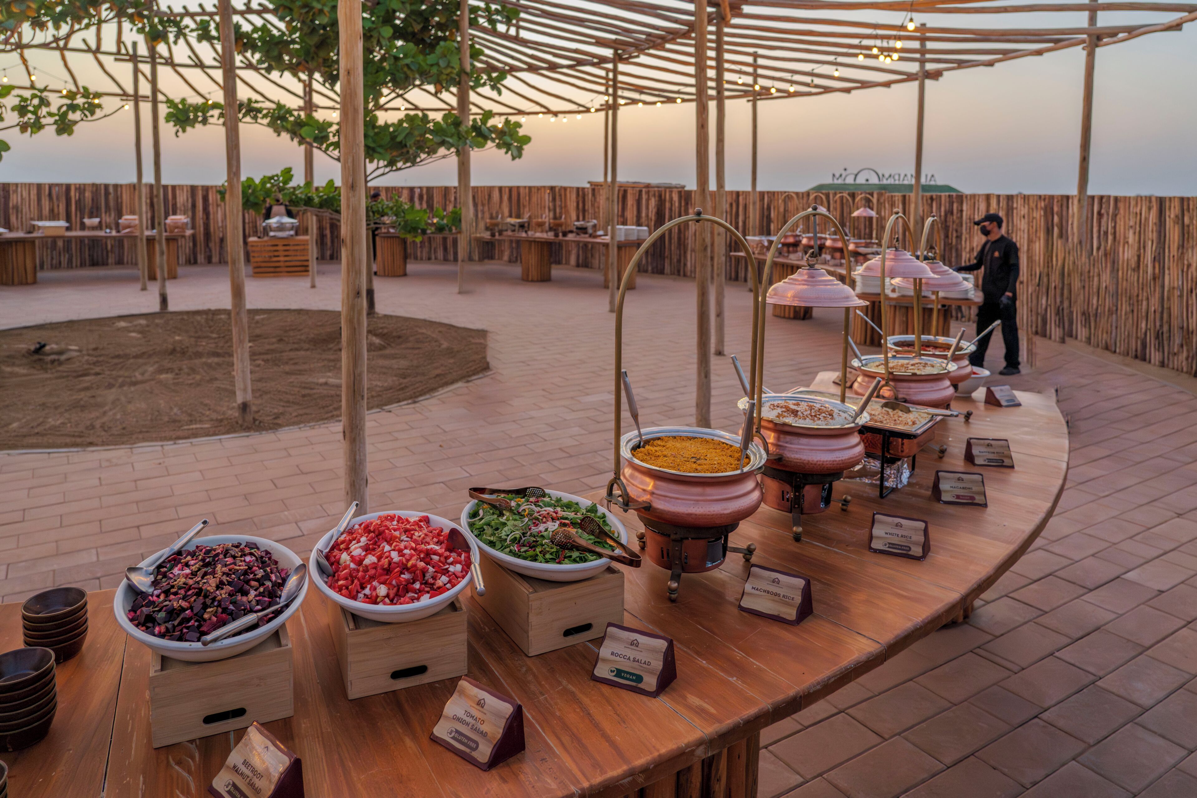 A buffet setup with various food containers and labels, a wooden table, and a person standing in the background.