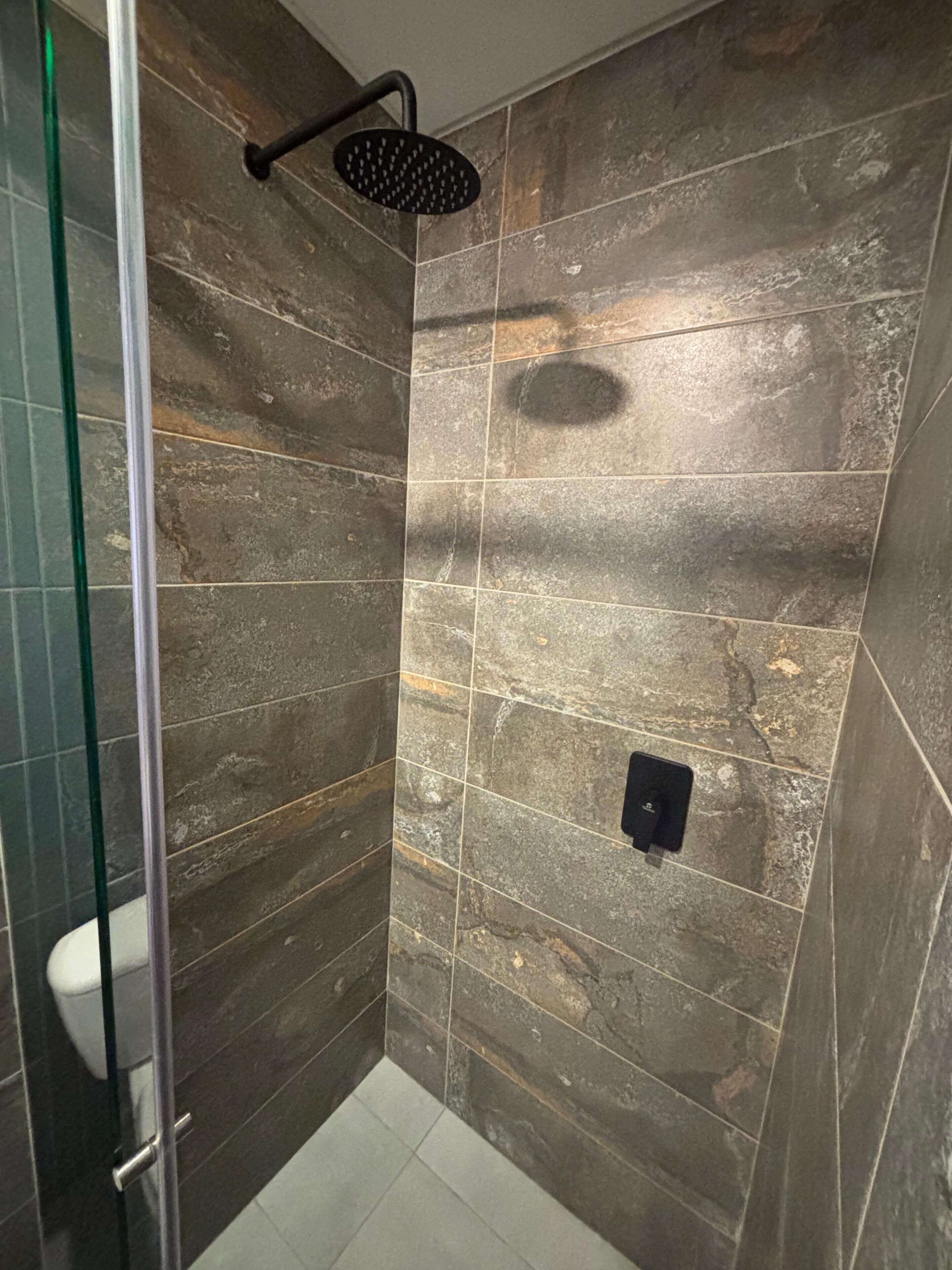 Un espace douche doté d’une porte en verre, d’un pommeau de douche à main et d’un mur carrelé à l’aspect pierre.