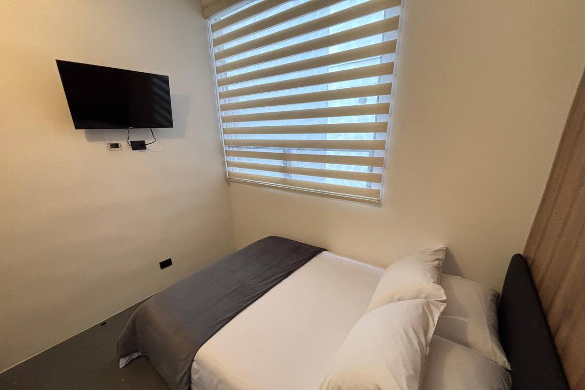 Ein kleines, ordentlich eingerichtetes Schlafzimmer mit einem Einzelbett, einem an der Wand befestigten Fernseher und einem Fenster mit Jalousien.