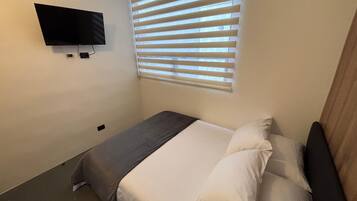 Ein kleines, ordentlich eingerichtetes Schlafzimmer mit einem Einzelbett, einem an der Wand befestigten Fernseher und einem Fenster mit Jalousien.