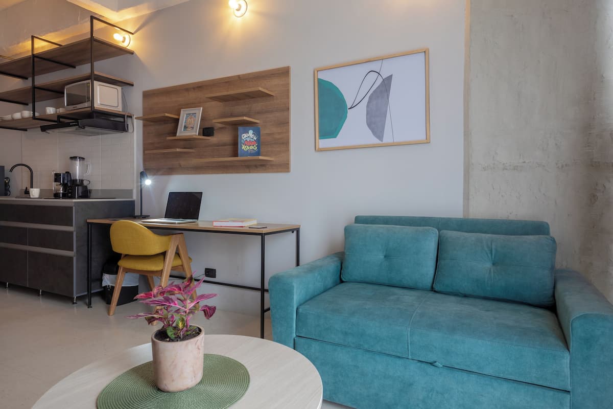 Ein modernes Wohnzimmer mit einem salbeengrünem Sofa, einem Essbereich mit einem Holztisch, einer Küche mit einer Mikrowelle und einem gerahmten abstrakten Gemälde an der Wand.