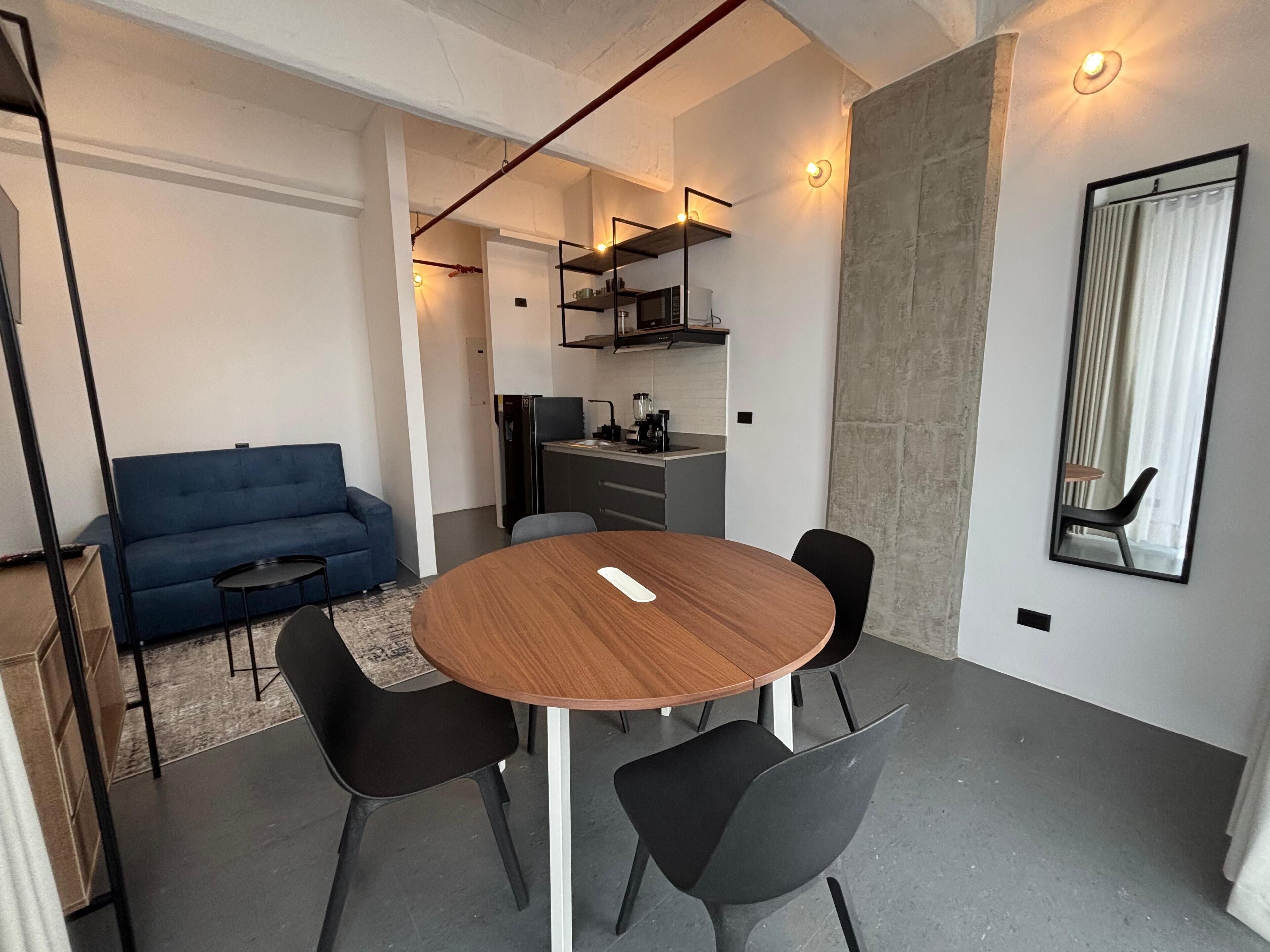 Una sala moderna con una mesa redonda de madera, sillas negras, un sofá azul y una cocina al fondo.