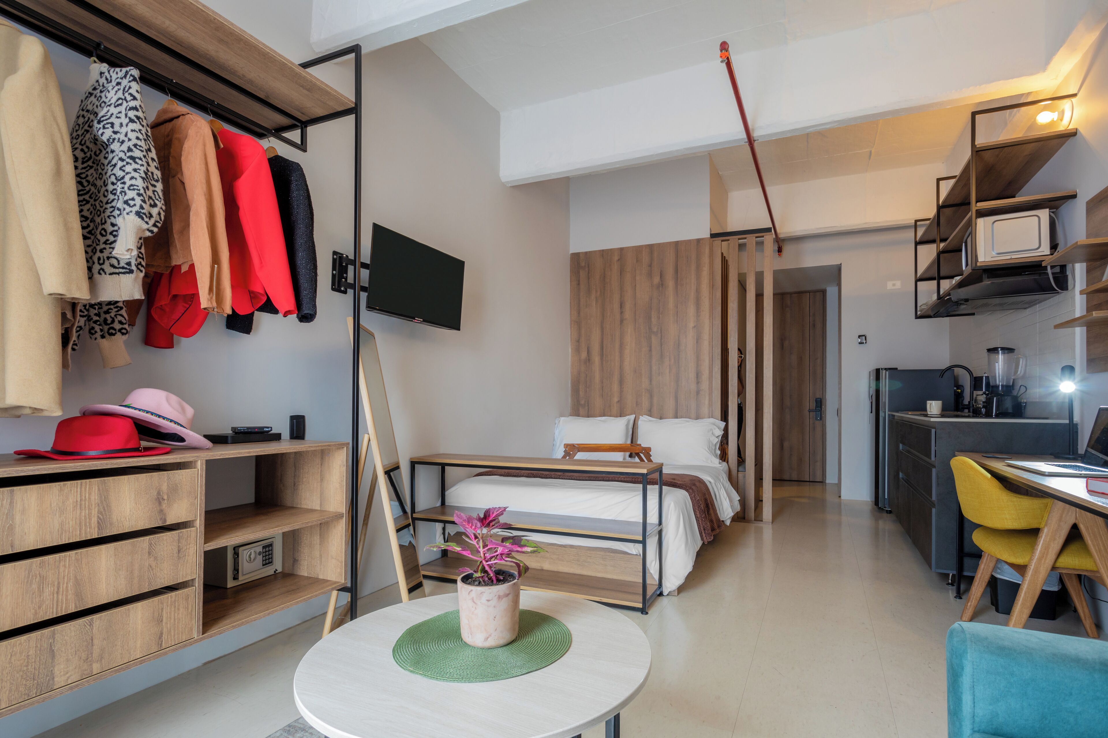 A compact, modern room with a bed, a desk, a chair, a TV, and a kitchenette.