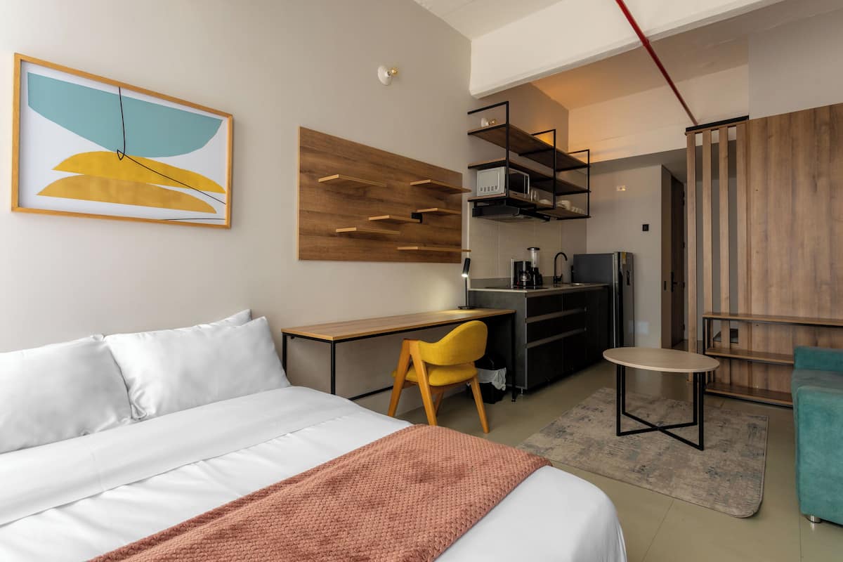 Ein modernes Hotelzimmer mit Bett, Schreibtisch, Stuhl, kleinem Tisch und einer Küchenzeile.
