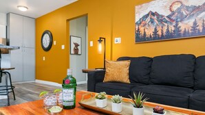 A living room with a black sofa, a wooden coffee table with a tray holding drinks and plants, and a painting of mountains on the wall. - Peterson Flats - Downtown Group Retreat (Fort Collins)