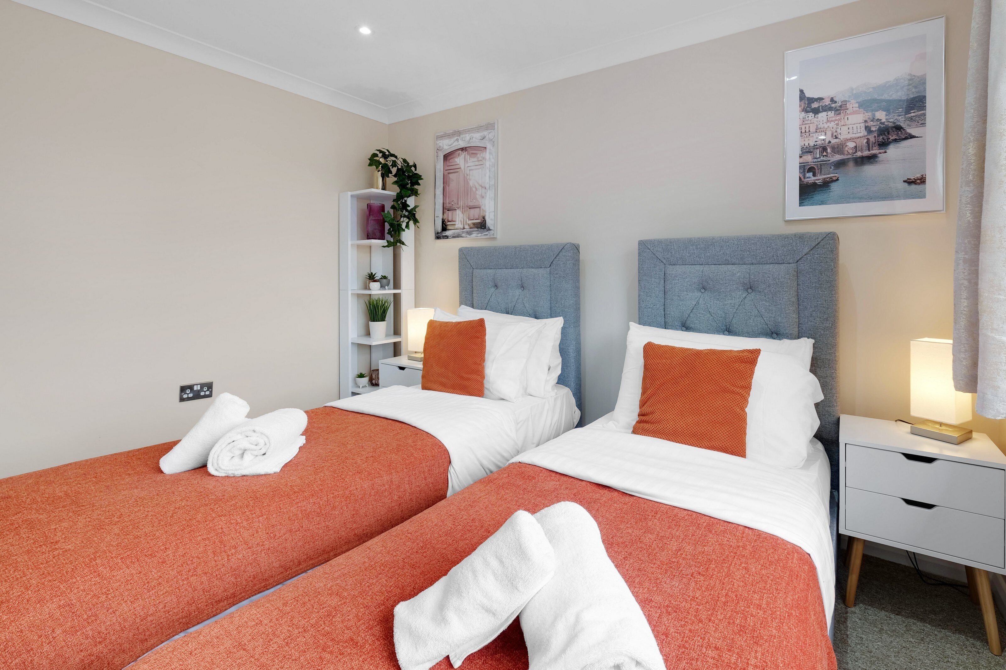 A hotel room with two beds, each with a grey headboard, white bedding, and orange pillows. There is a nightstand with a lamp and a small shelf with plants.