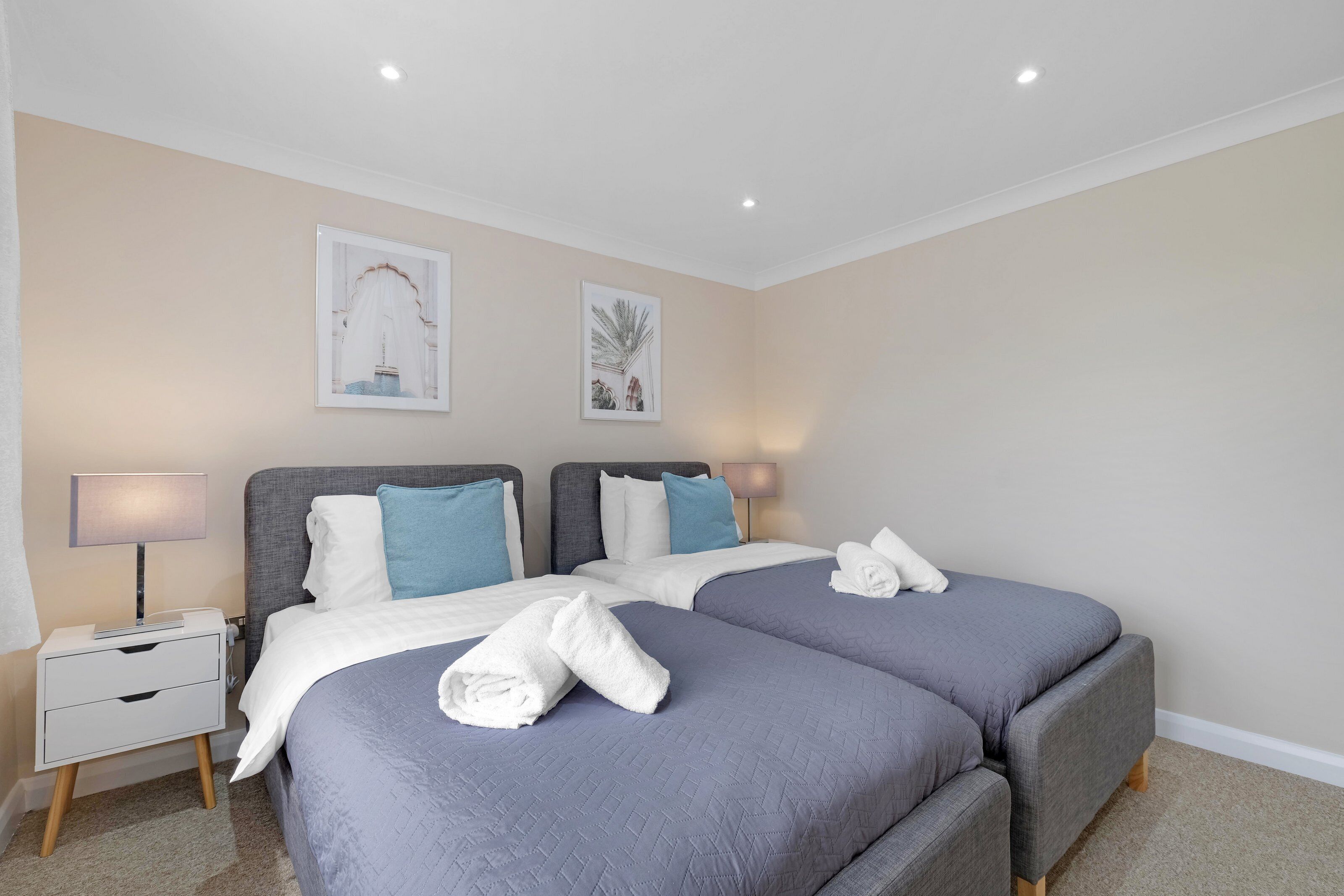 Habitación doble con dos camas, cada una con cabecero gris y ropa de cama blanca. Hay dos mesitas de noche con lámparas, y se exhibe arte en las paredes.