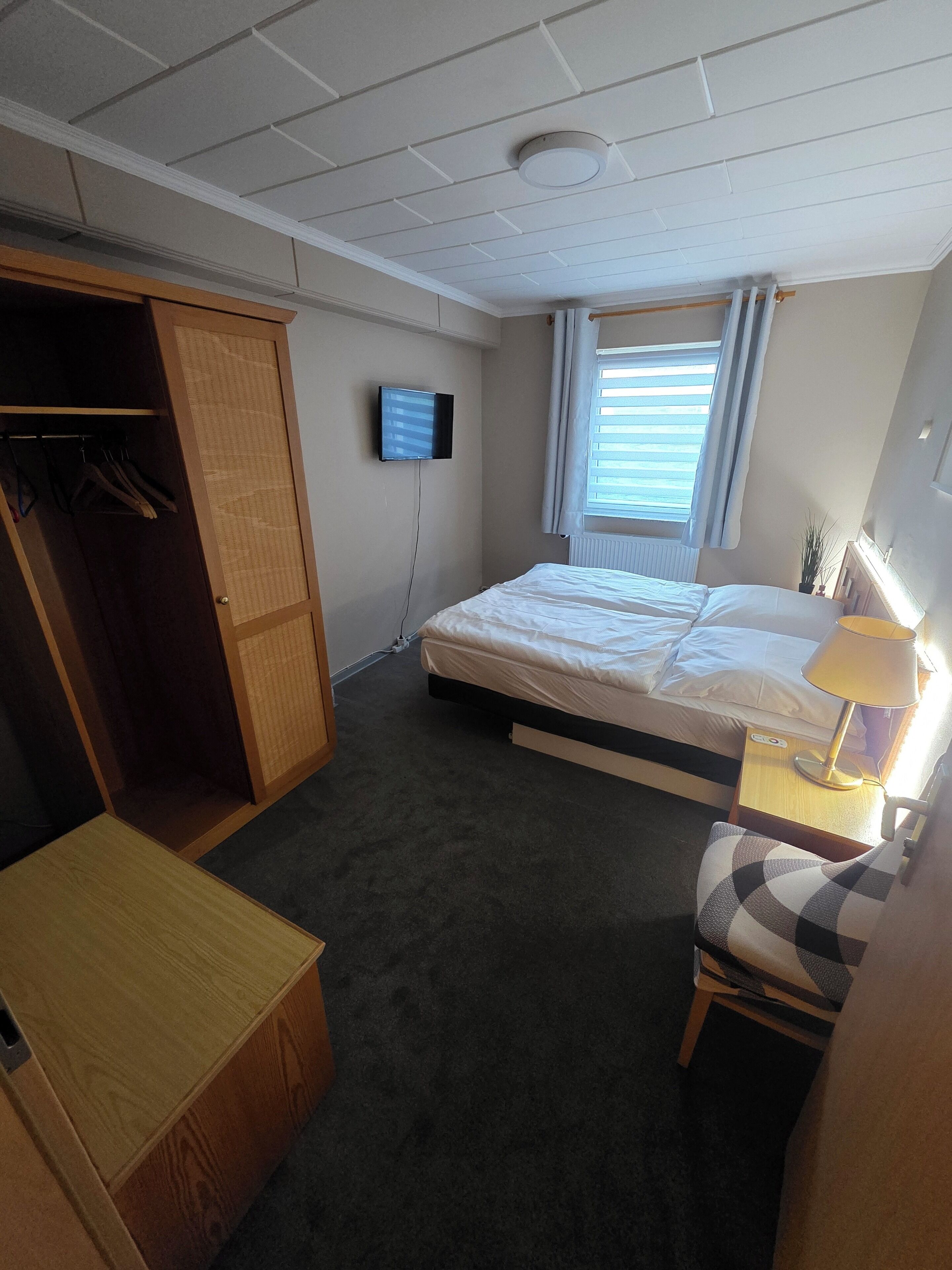 Quarto de hotel com cama, televisão na parede, guarda-roupa, secretária com candeeiro e janela com persianas.