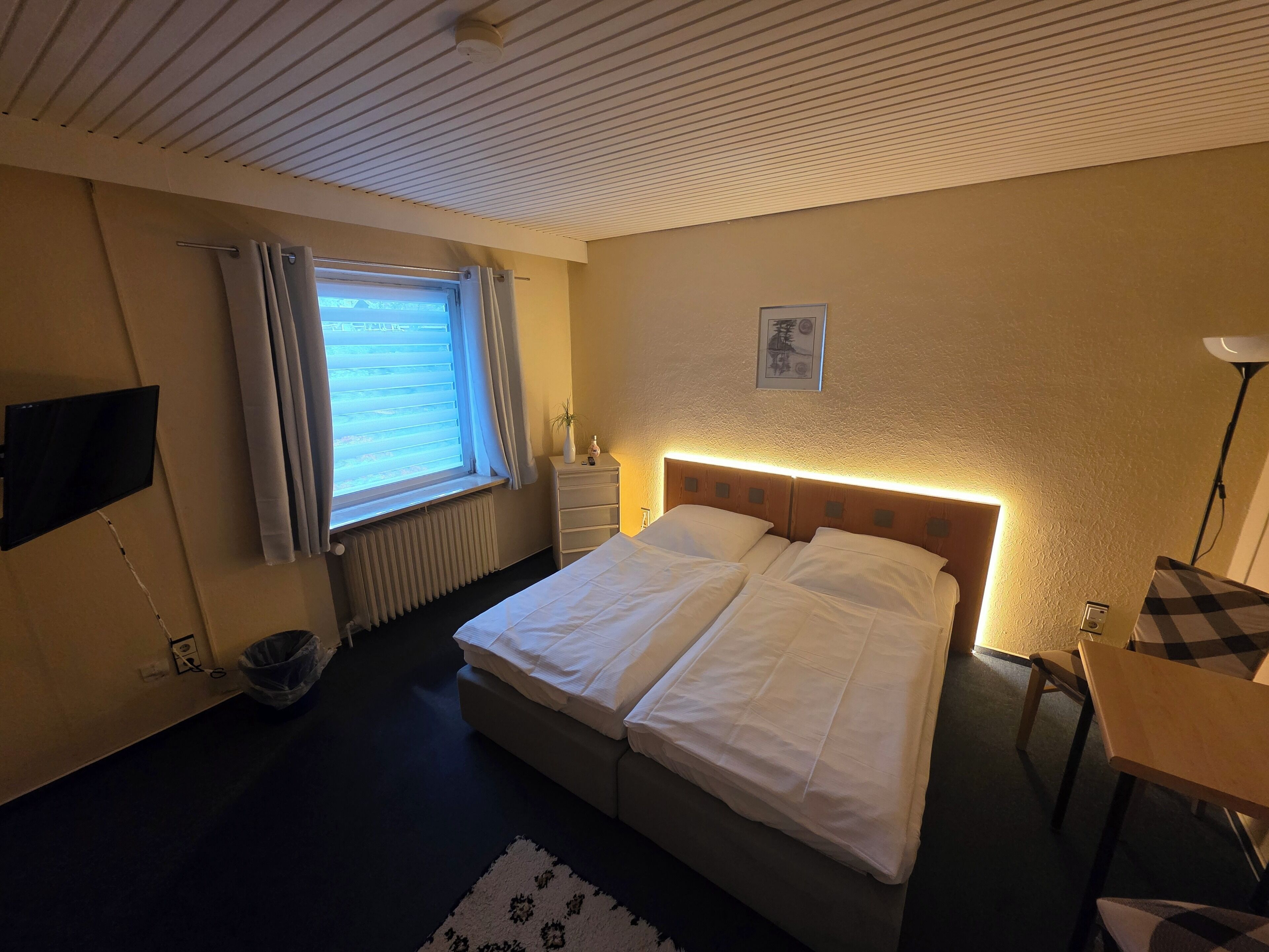 Quarto de hotel com cama, mesa de cabeceira, abajur, janela com cortinas, radiador e televisão presa à parede.
