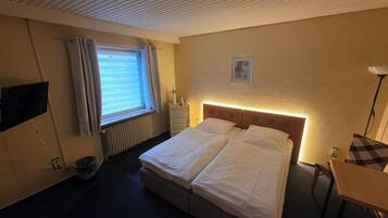 Quarto de hotel com cama, mesa de cabeceira, abajur, janela com cortinas, radiador e televisão presa à parede.