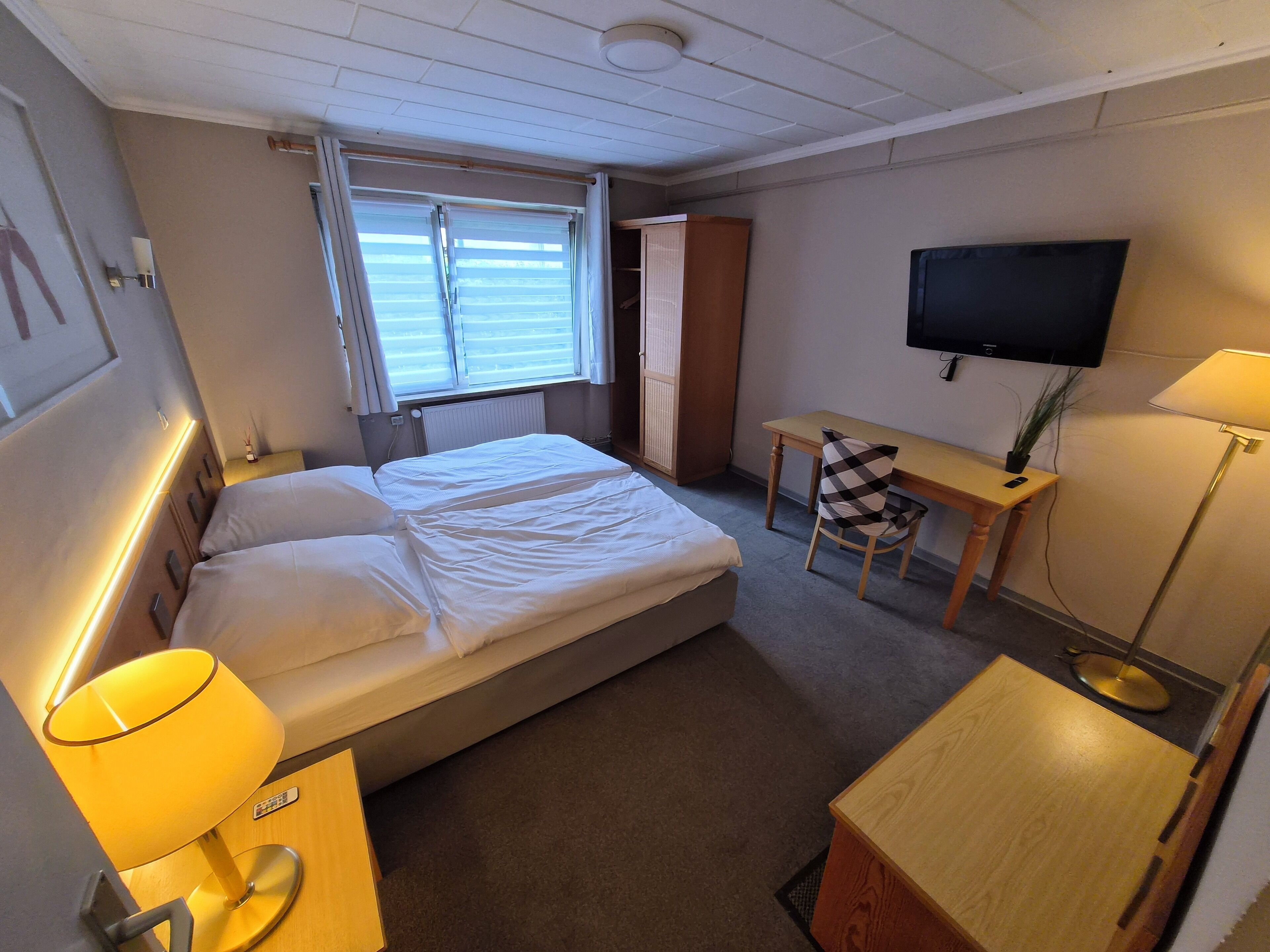 Ein Hotelzimmer mit Bett, Nachttisch, Lampe, Schreibtisch mit Stuhl, Fernseher und einem Fenster mit Jalousien.
