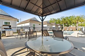 A covered patio area with a glass table and chairs, a barbecue grill, and a swimming pool in the background. - Cozy Cottage 4 of 6 with Pool Views| 2BR | Pool (Port St. Joe)