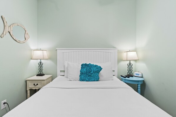 A neatly made bed with white linens and a teal accent pillow, flanked by two bedside tables with lamps and a radio, in a room with light green walls and a wooden fish-shaped decor.