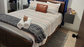 Una cama bien tendida con ropa de cama a rayas, un cojín de cuero marrón y toallas enrolladas. Hay una mesita de noche pequeña con una lámpara y un letrero que dice 