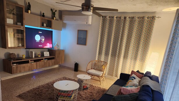 Una sala de estar con televisor, mueble de madera para equipo audiovisual, dos mesas redondas, un sofá y una cortina estampada.