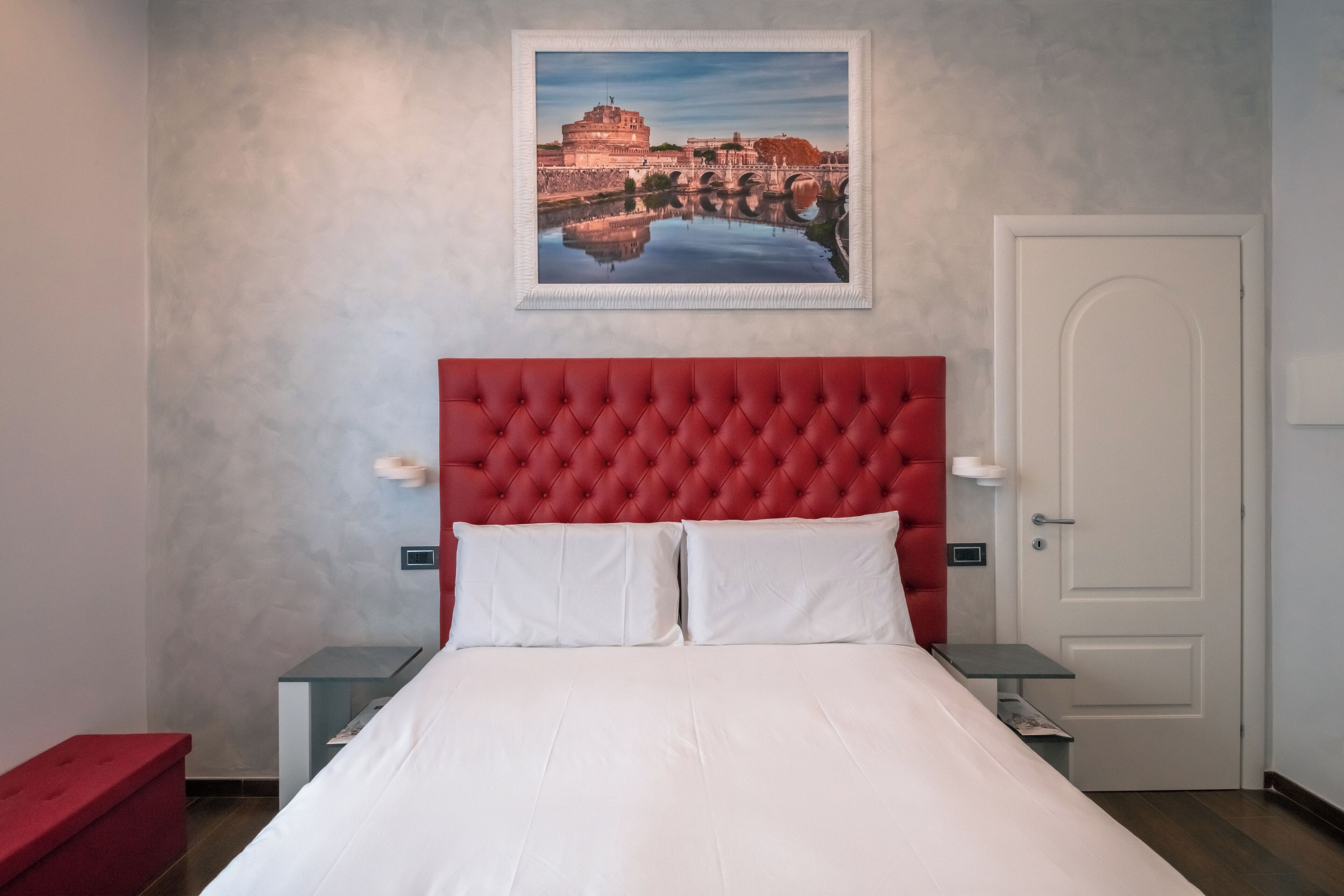 A hotel room with a red tufted headboard, white bedding, a framed picture of a historical building, and a white door.