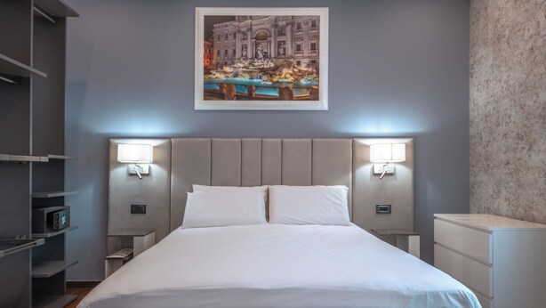 Une chambre d’hôtel moderne dotée d’un grand lit, de deux tables de chevet, d’une petite table et d’une œuvre d’art fixée au mur représentant une fontaine historique.