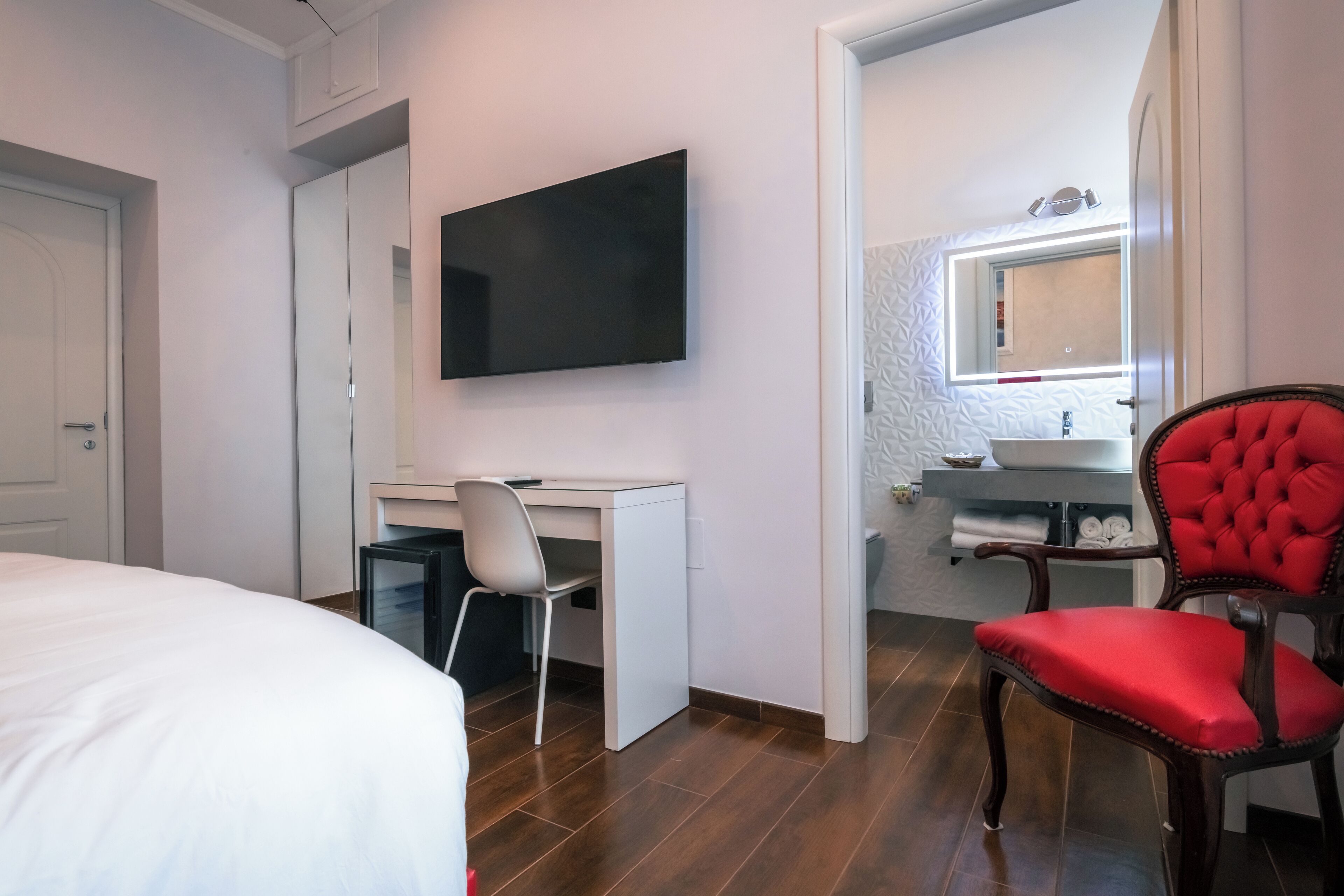 Une chambre d’hôtel comprenant un lit, un bureau avec une chaise, une télévision, un fauteuil rouge rembourré et une salle de bain avec un miroir et un lavabo.