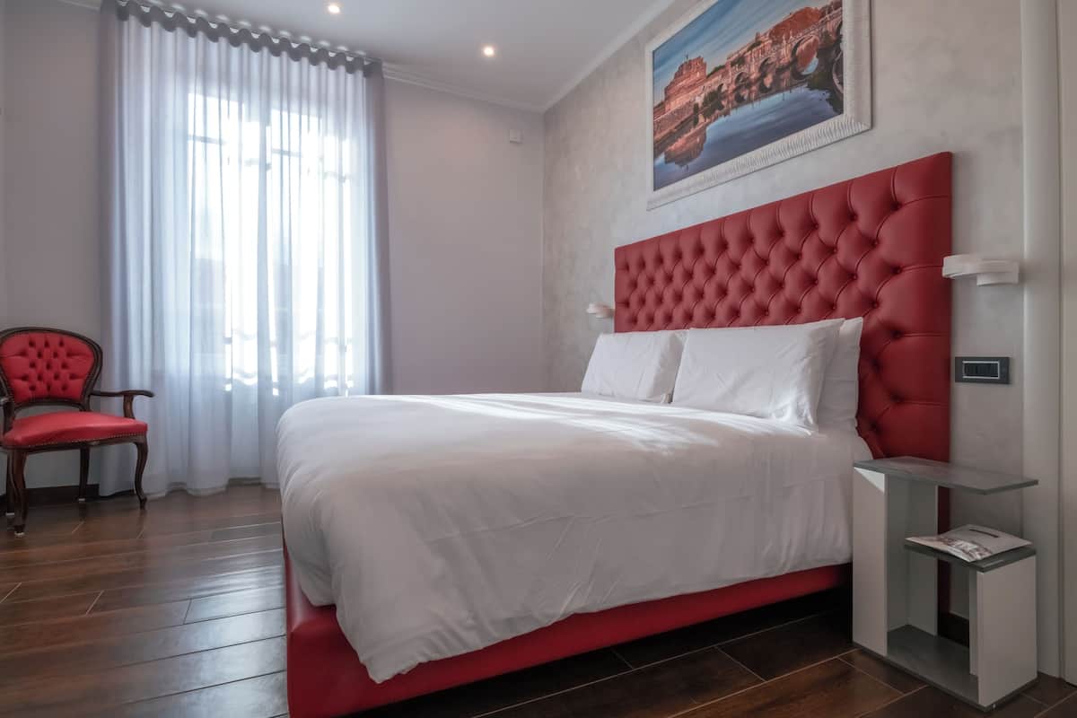 A bedroom with a red upholstered headboard, a white bedspread, a wooden chair, and a painting on the wall.